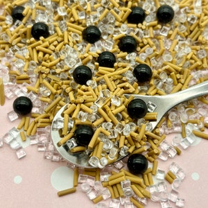 May include: A close-up of a spoon filled with brown and clear sprinkles, black sprinkles, and black balls. The sprinkles are scattered on a pink surface with white dots.