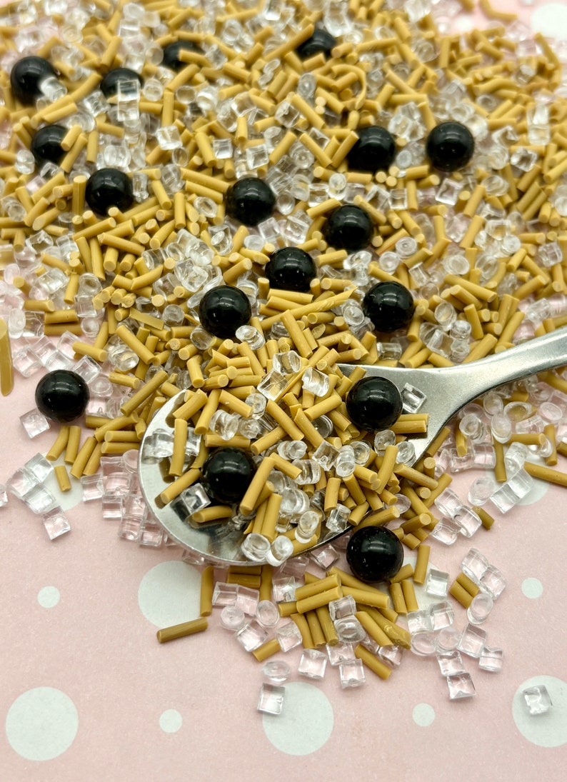 May include: A close-up of a spoon filled with brown and clear sprinkles, black sprinkles, and black balls. The sprinkles are scattered on a pink surface with white dots.