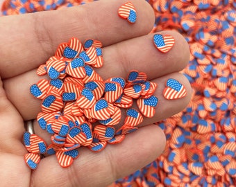Op de afbeelding: Een close-up van een hand die een stapel kleine rode, witte en blauwe hartvormige sprinkles met een Amerikaans vlagontwerp vasthoudt.