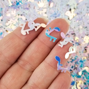 May include: A hand holding iridescent holographic glitter in the shape of music notes. The glitter is a mix of white, blue, and pink.