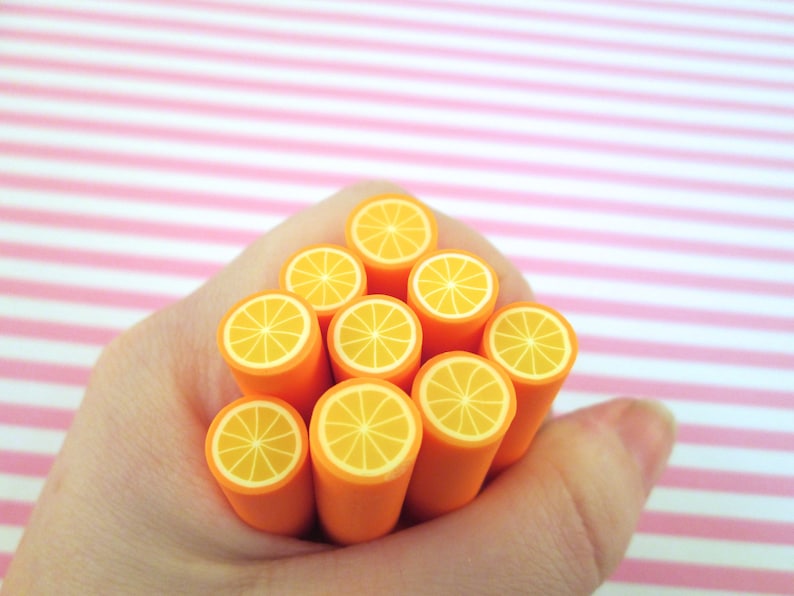May include: A hand holding a stack of orange polymer clay canes with a slice of orange design. The canes are arranged in a pyramid shape.