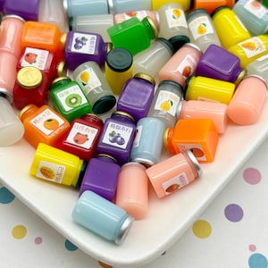 May include: A collection of miniature, colorful juice bottles with silver lids, arranged on a white plate. The bottles are various colors, including purple, orange, yellow, and pink, and feature labels with fruit illustrations and Chinese characters. The plate sits on a polka dot surface.