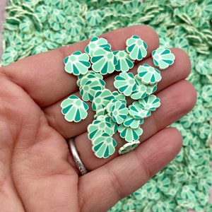 May include: A close-up of a hand holding a pile of green and white flower-shaped resin embellishments. The embellishments are perfect for adding a touch of whimsy to crafts and projects.