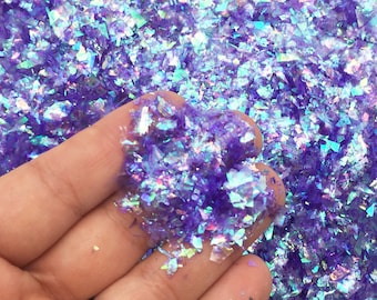 May include: Close-up of iridescent purple glitter flakes. The flakes are irregularly shaped and reflect multiple colors, including blue and green. The glitter is held in a hand, with the background filled with more of the same glitter.