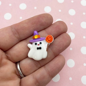 May include: A white ghost wearing a purple witch hat with a yellow buckle and holding a lollipop. The ghost has black eyes and a black bow tie.