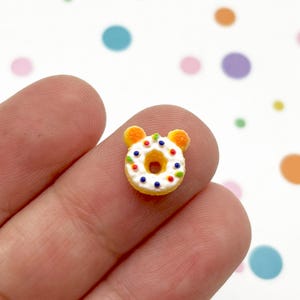 Puede incluir: Un mini donut amarillo con glaseado blanco y chispas de colores. El donut tiene dos pequeñas orejas de oso naranjas en la parte superior. El donut se sostiene entre dos dedos sobre un fondo blanco con lunares de colores.