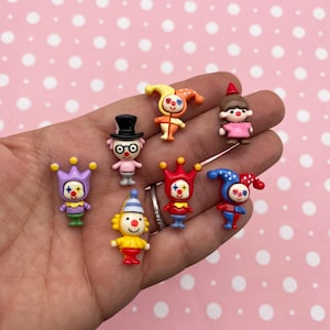 Puede incluir: Una colección de pequeñas figuras de payasos de colores. Cada figura tiene un diseño único, con diferentes sombreros, trajes y expresiones faciales. Los payasos son de varios colores, incluyendo rojo, amarillo, azul y morado. El fondo es una superficie rosa con lunares blancos.