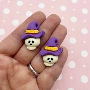 Può includere: Un paio di orecchini a forma di teschio di strega viola e gialli. I teschi indossano cappelli da strega viola con fasce gialle.
