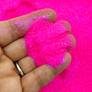 May include: A close-up of a hand holding a pile of bright pink glitter with iridescent shimmer. The glitter is fine and sparkly, and it is perfect for crafting and decorating.