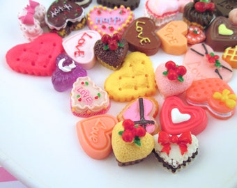 May include: A collection of heart-shaped decorative items in various colors and designs. Some have the word "Love" or "Happy" on them. Others are decorated with bows, cherries, and other embellishments. The items appear to be made of resin or a similar material.