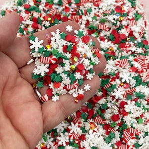 May include: A close-up of a hand holding a variety of Christmas sprinkles. The sprinkles are red, green, and white and include snowflakes, stars, candy canes, and small red circles.