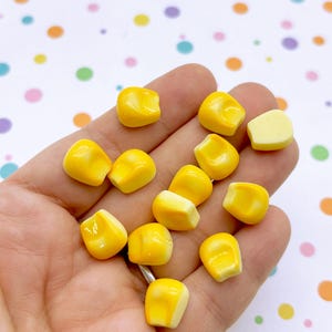 May include: A handful of small, bright yellow corn kernels. The kernels have a glossy finish. The background is a white surface with colourful polka dots.