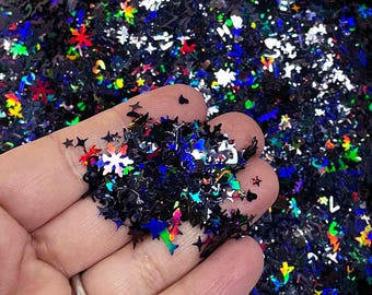 Pode incluir: Um close-up de uma mão segurando uma variedade de glitter iridescente em preto, azul, verde, vermelho e prata. O glitter tem a forma de estrelas, flocos de neve e outras formas geométricas.