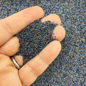 May include: A hand holding a pile of small, iridescent beads. The beads are primarily dark blue with hints of gold and purple. A silver ring with small stones is visible on the finger.