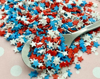 Op de afbeelding: Een zilveren lepel gevuld met rode, witte en blauwe stervormige sprinkles. De sprinkles zijn verspreid over een roze oppervlak met witte stippen.