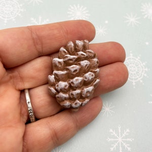 May include: A small, brown pine cone with white paint accents. The pine cone is about 2 inches tall and is a natural, rustic decoration.