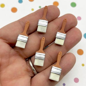 May include: Six miniature paintbrushes are shown, each with a brown handle, silver ferrule, and white bristles. These small paintbrushes are likely for crafts or decorative use. The background is white with colourful polka dots.