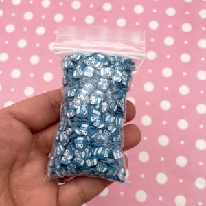 K&ouml;nnte beinhalten: Ein durchsichtiger Plastikbeutel gef&uuml;llt mit blauen und wei&szlig;en Milchpackungs-f&ouml;rmigen Streuseln. Die Streusel haben ein Comic-Gesicht mit Augen und einem L&auml;cheln. Der Beutel wird in einer Hand gehalten.