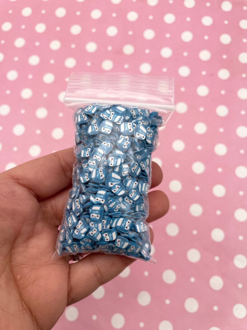 K&ouml;nnte beinhalten: Ein durchsichtiger Plastikbeutel gef&uuml;llt mit blauen und wei&szlig;en Milchpackungs-f&ouml;rmigen Streuseln. Die Streusel haben ein Comic-Gesicht mit Augen und einem L&auml;cheln. Der Beutel wird in einer Hand gehalten.