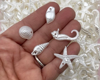 May include: A collection of white pearl-like sea-themed charms, including a starfish, seashell, and a seahorse. The charms are displayed on a hand.