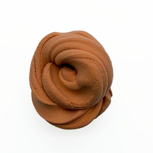 May include: A close-up shot of a swirl of brown slime, resembling a rose. The slime has a textured, matte finish and is set against a white background. The color is a warm, earthy tone.