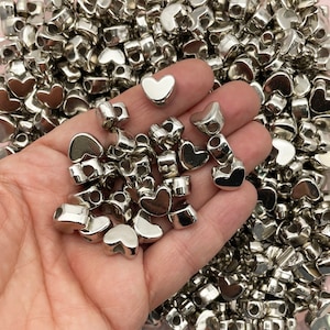 May include: A hand holding a large quantity of silver heart-shaped beads with a hole through the center. The beads are perfect for jewelry making or crafting.