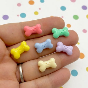 May include: Six pastel-colored plastic dog bone charms with a smiley face on each. The charms are in the colors pink, green, light blue, purple, yellow, and cream.