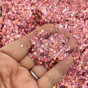 May include: A handful of pink glitter in various shapes and sizes, including hexagons and smaller particles. The glitter has a shimmering, iridescent quality, reflecting light with shades of pink, orange, and purple. The glitter is held in a hand.