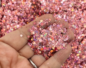 May include: A handful of pink glitter in various shapes and sizes, including hexagons and smaller particles. The glitter has a shimmering, iridescent quality, reflecting light with shades of pink, orange, and purple. The glitter is held in a hand.