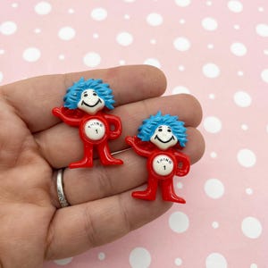 Könnte beinhalten: Zwei rote Thing 1 und Thing 2 Figuren mit blauen Haaren und weißen Gesichtern. Jede Figur hat die Nummer 1 auf der Brust. Die Figuren werden in einer Hand vor einem rosa Hintergrund mit weißen Punkten gehalten.