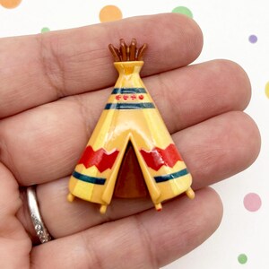 May include: A miniature, yellow teepee with red and blue decorative accents. The small, three-dimensional structure has a brown top and is likely a decorative item or toy.