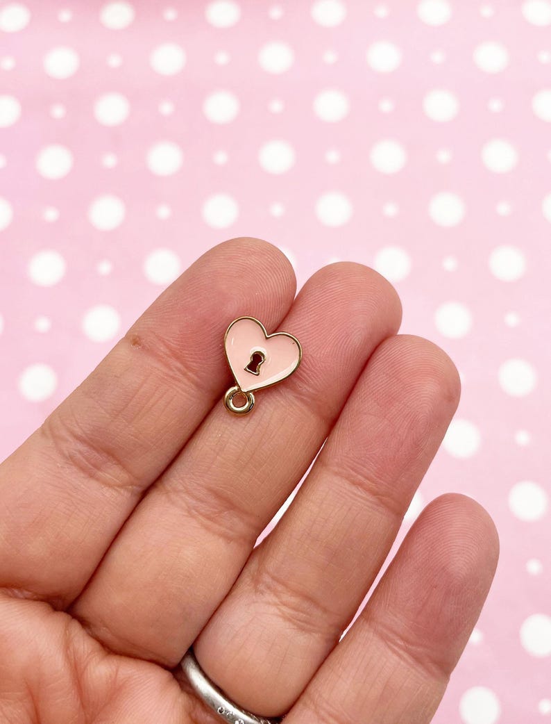 May include: A small, pink heart-shaped charm with a keyhole design. The charm has a gold-colored border and a small loop at the top for attaching to jewelry or other crafts. The background is a light pink color with white polka dots.