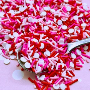May include: A close-up of a spoon filled with red, pink, and white sprinkles. The sprinkles are a mix of small round sprinkles and long, thin sprinkles.