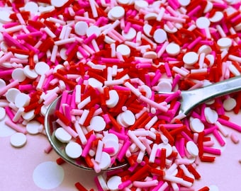 May include: A close-up of a spoon filled with red, pink, and white sprinkles. The sprinkles are a mix of small round sprinkles and long, thin sprinkles.