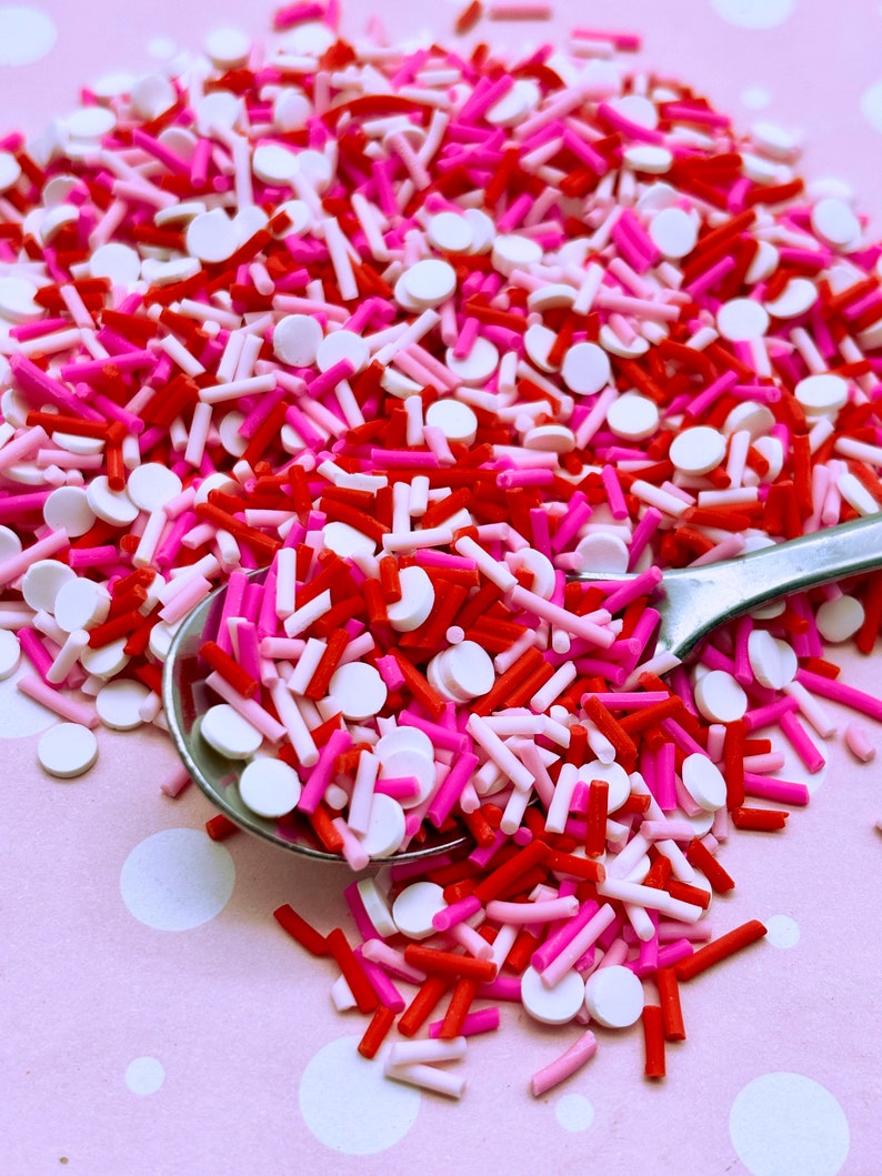 May include: A close-up of a spoon filled with red, pink, and white sprinkles. The sprinkles are a mix of small round sprinkles and long, thin sprinkles.