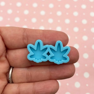 Puede incluir: Un molde de silicona azul claro con dos formas de hojas de cannabis. El molde está diseñado para hacer aretes de resina o arcilla polimérica.