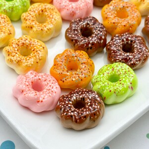 May include: A collection of colorful, miniature donuts in various colors, including pink, yellow, orange, green, and brown. Each donut has a hole in the center and is decorated with sprinkles or frosting. The donuts are arranged on a white plate.