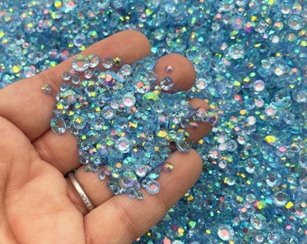 May include: A close-up of a hand holding a large amount of iridescent light blue rhinestones. The rhinestones are scattered and have a variety of sizes.