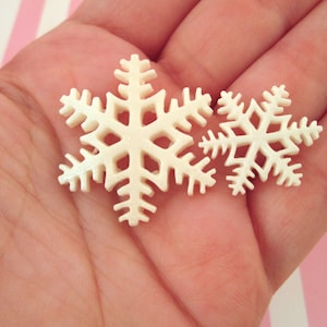 May include: Two white snowflake ornaments, one larger than the other, are held in a hand against a pink and white striped background.