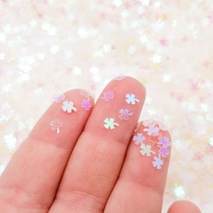以下が含まれることがあります： 手の中に、虹色、透明、ピンクの四つ葉のクローバー型のキラキラしたものが握られています。キラキラしたものは白い表面に散らばっています。