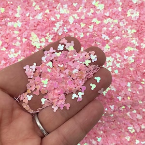 May include: Pink iridescent glitter in the shape of Mickey Mouse heads. The glitter is scattered on a surface and a hand is holding a small pile of the glitter.