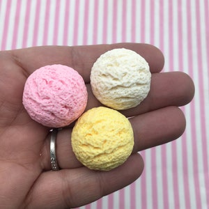 Pu&ograve; includere: Tre palline di gelato rosa, giallo e bianco, tenute in mano. Le palline hanno una superficie strutturata, che ricorda il gelato vero. Lo sfondo &egrave; un motivo a strisce rosa e bianche.