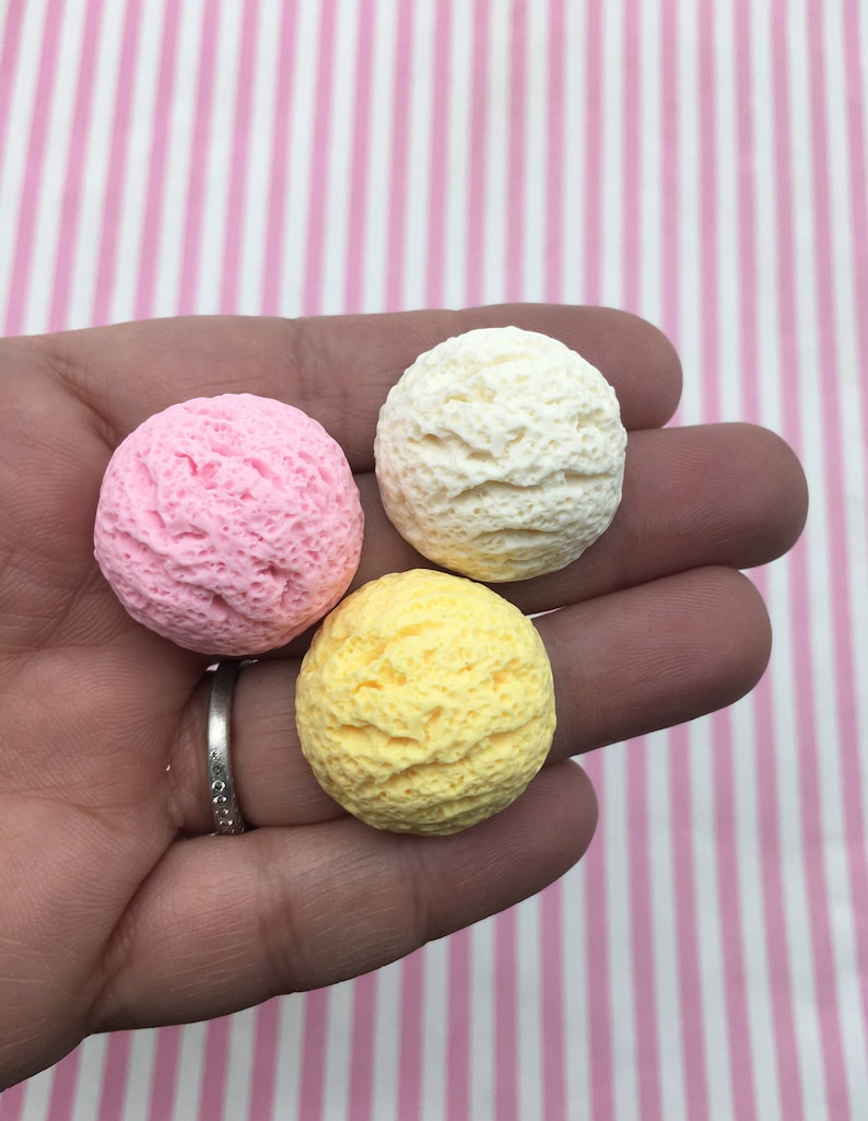 Pu&ograve; includere: Tre palline di gelato rosa, giallo e bianco, tenute in mano. Le palline hanno una superficie strutturata, che ricorda il gelato vero. Lo sfondo &egrave; un motivo a strisce rosa e bianche.