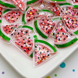 Op de afbeelding: Een verzameling watermeloen-vormige bedels. Elke bedel heeft een doorzichtige plastic behuizing gevuld met rode, roze en glinsterende stervormige confetti. De watermeloenplakken hebben groene schillen, wit vruchtvlees en zwarte zaaddetails.