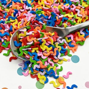 May include: A spoon filled with colorful, wavy sprinkles. The sprinkles are in a variety of colors, including red, orange, yellow, green, blue, and purple. The sprinkles are scattered on a white surface with colorful polka dots.