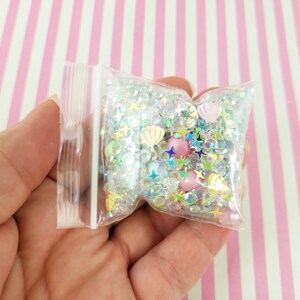 May include: A clear plastic bag filled with iridescent glitter, small stars, and seashells. The bag is held in a hand.