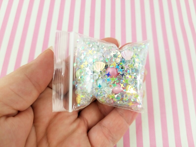 May include: A clear plastic bag filled with iridescent glitter, small stars, and seashells. The bag is held in a hand.