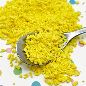 May include: A close-up of a spoon scooping up yellow sprinkles. The sprinkles are scattered on a white surface with colorful polka dots.