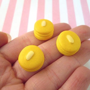 May include: Three yellow miniature pancake charms with a white dollop of icing on top. The charms are on a hand.