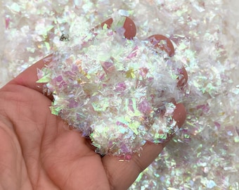 Könnte beinhalten: Eine Handvoll schimmernder weißer Glitzerflocken. Der Glitzer besteht aus kleinen, unregelmäßigen Plastikstücken, die das Licht in einem Regenbogen von Farben reflektieren.